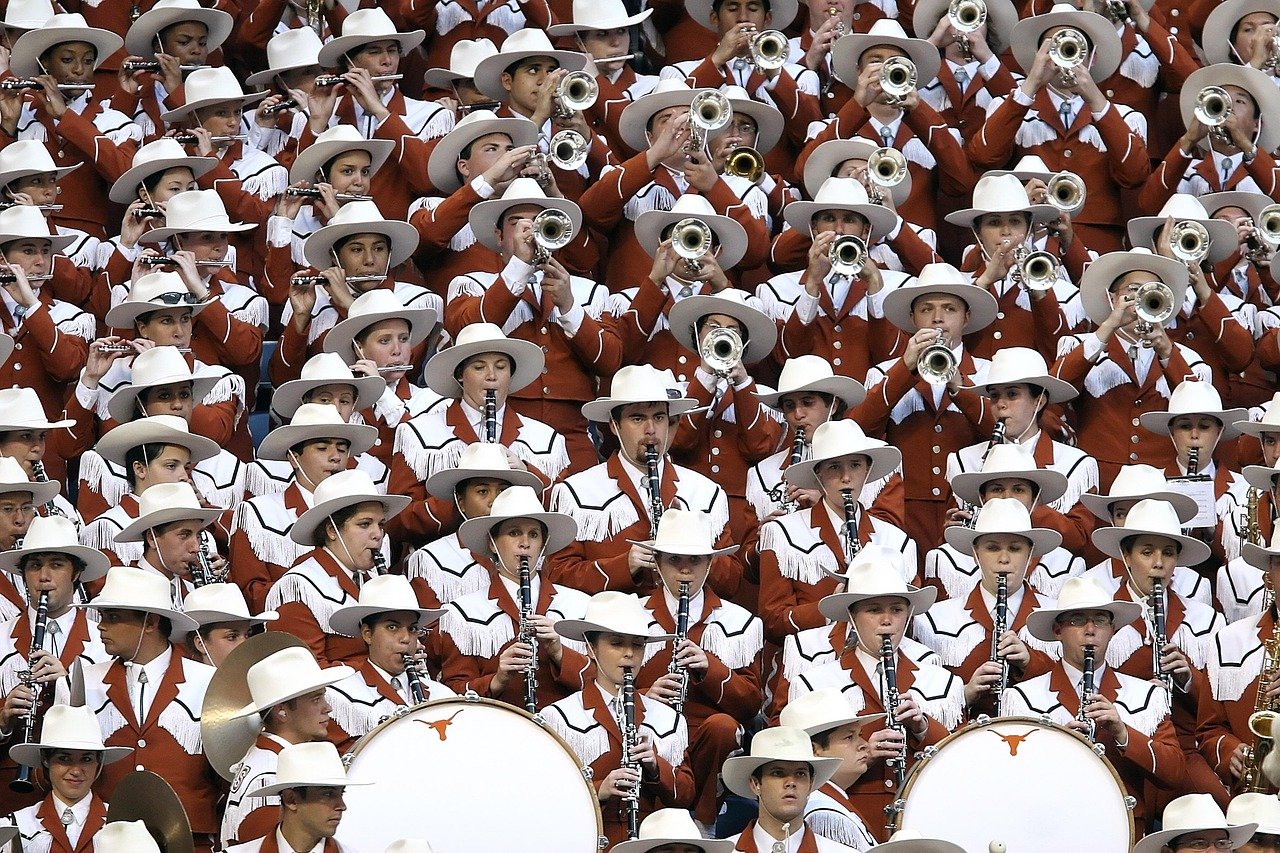 College marching band members in brown uniforms and white cowboy hats performing with brass instruments at stadium